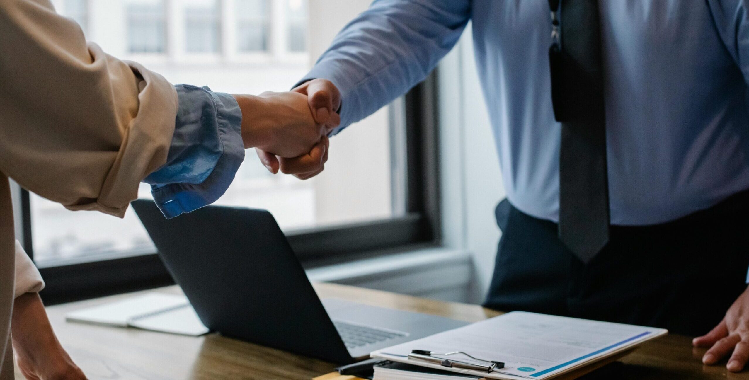 Crop unrecognizable coworkers in formal wear standing at table with laptop and documents while greeting each other before meeting